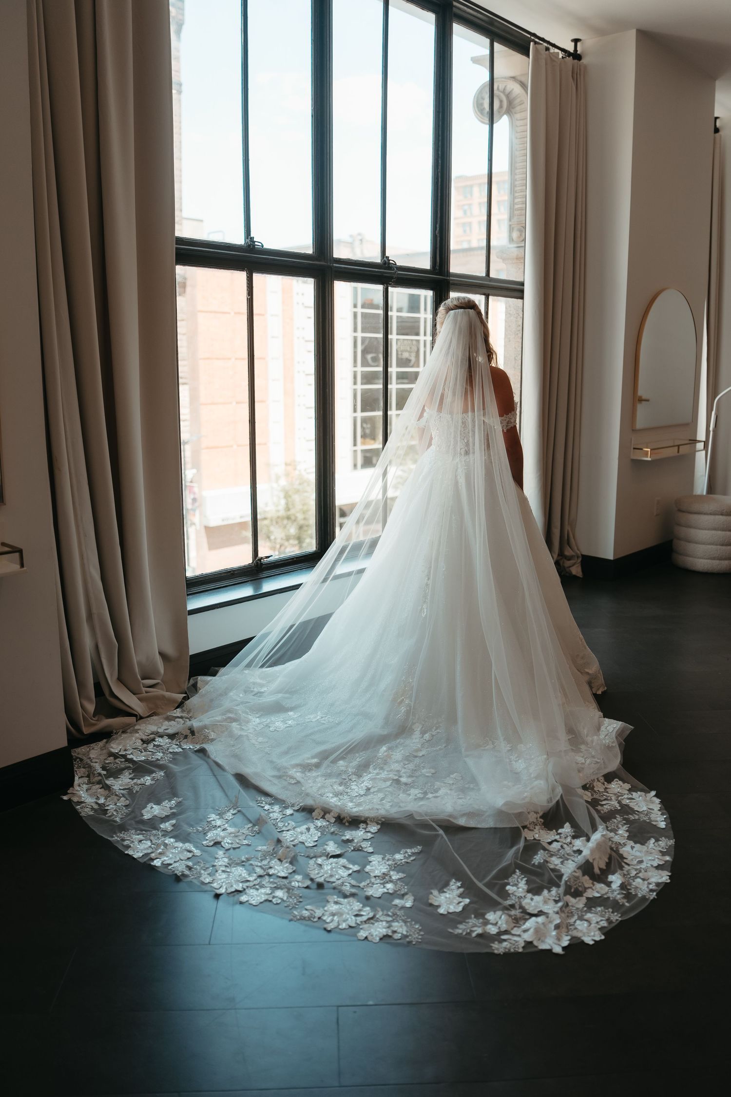 Bride in a white ball gown with a floral lace cathedral veil stands facing large windows in an elegant room.