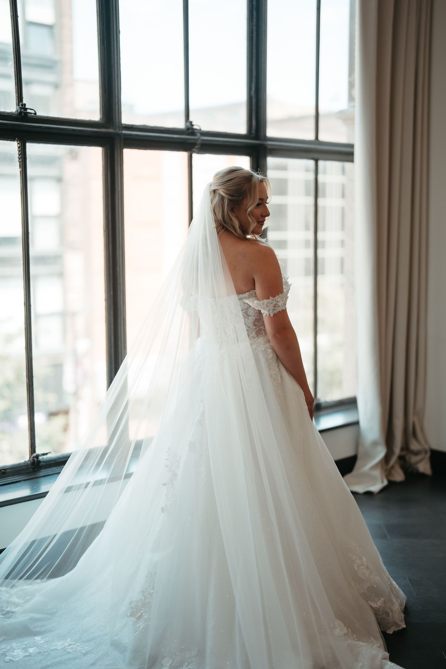 Smiling bride in an off-shoulder white ball gown and long veil poses by tall windows in a bright room.
