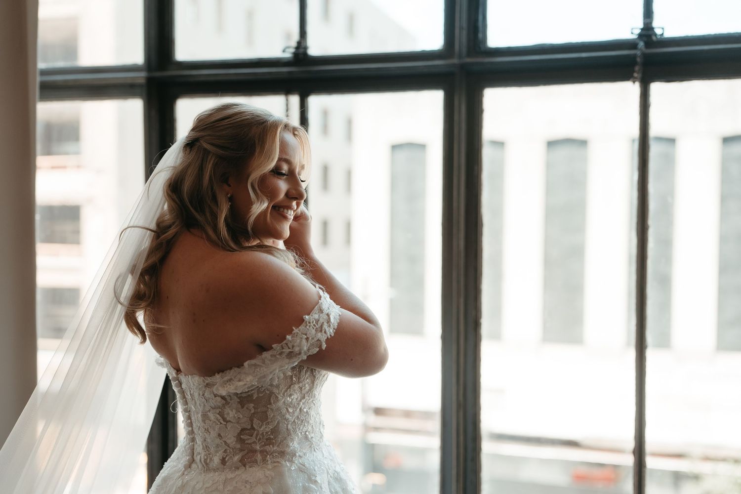 Bride in lace off-shoulder wedding gown and veil smiling by large window in urban setting.
