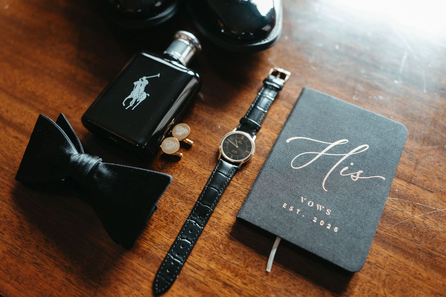 Groom wedding flat lay featuring Polo Ralph Lauren cologne, black bow tie, cufflinks, watch, and a His Vows journal on wood.