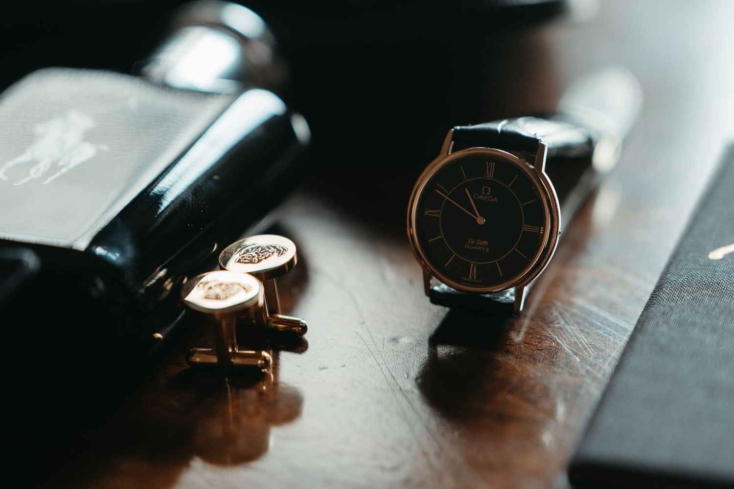 Classic wristwatch with black leather strap beside gold cufflinks and cologne bottle on dark wooden surface.
