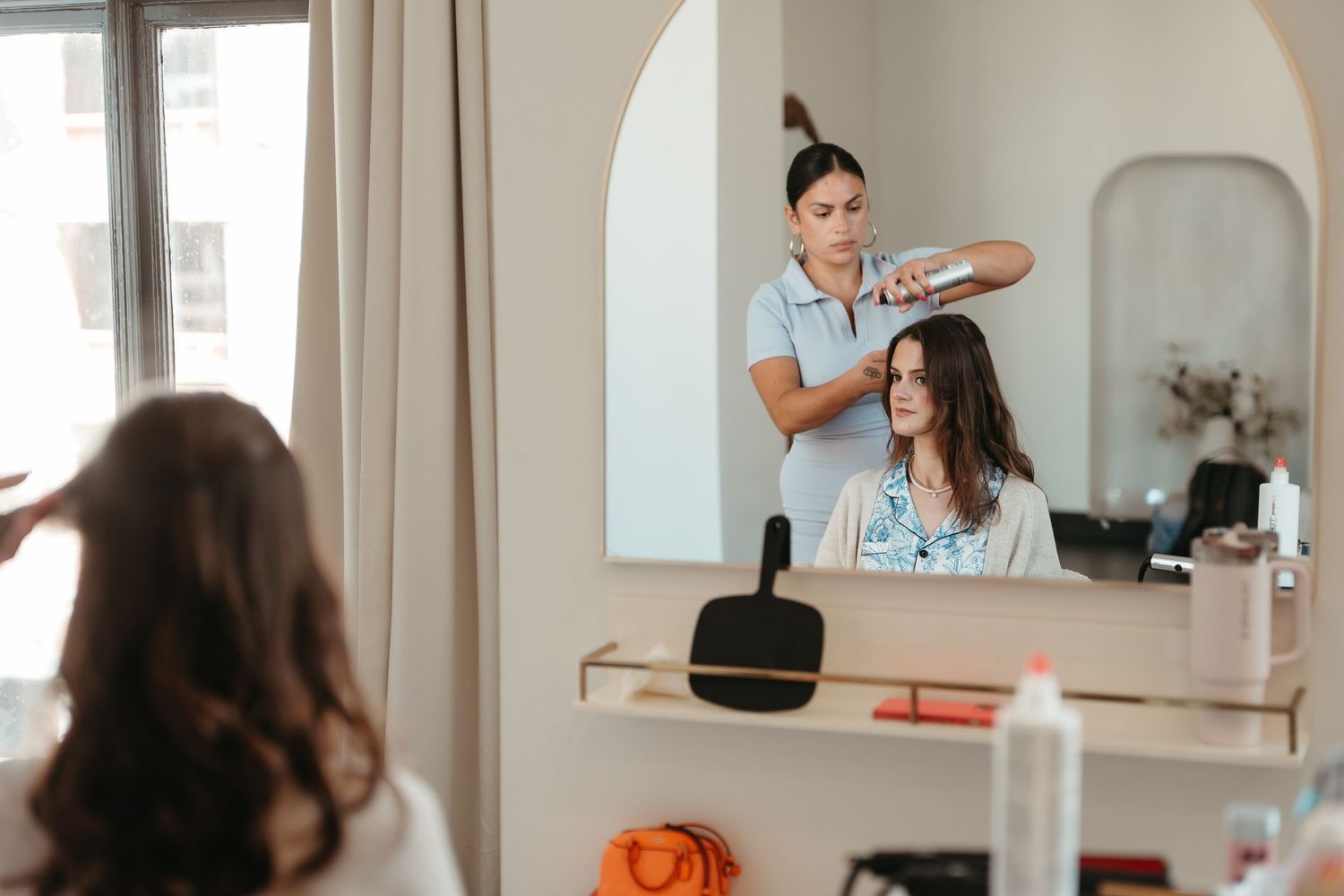 Hairstylist blow-drying a brunette client's hair in a modern salon, reflected in an arched mirror.