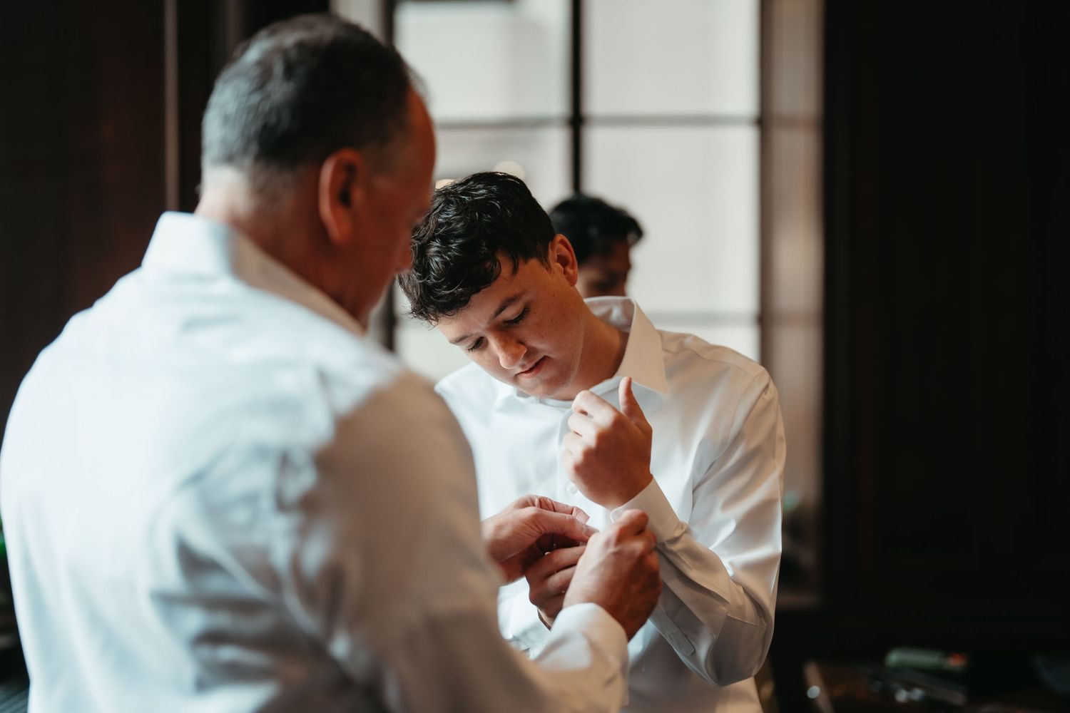 Two men in white dress shirts adjusting cufflinks indoors, preparing for a formal wedding event.