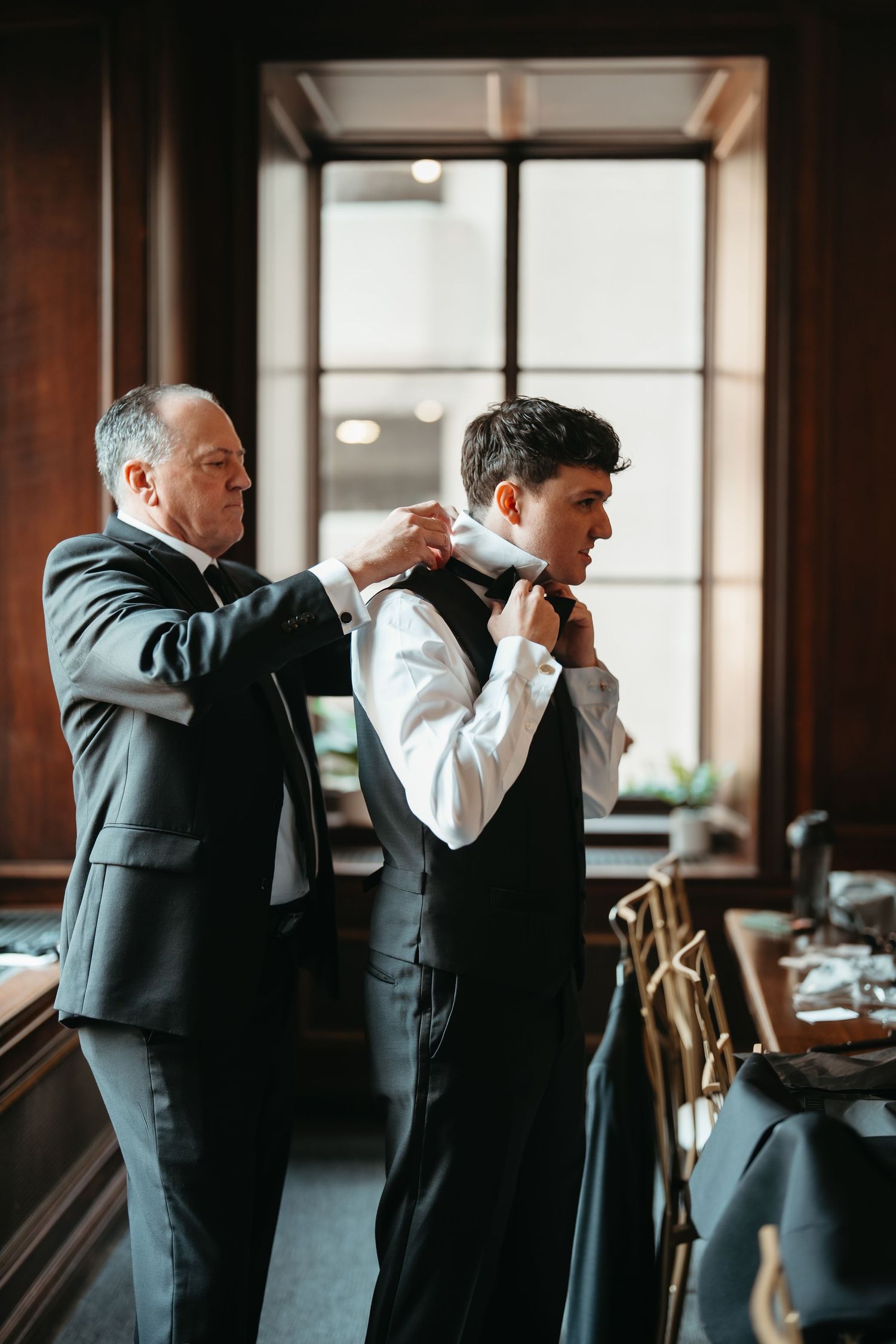 An older man helps a young groom adjust his bow tie in an elegant wood-paneled room before a wedding ceremony.