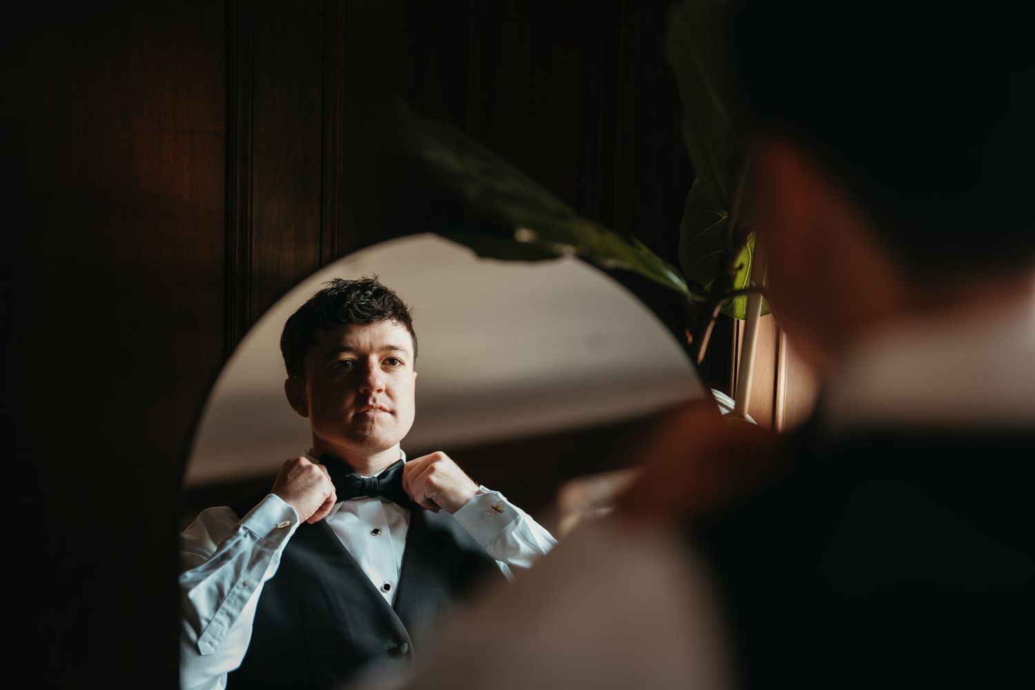 Groom adjusting bow tie in mirror reflection, wearing tuxedo while getting ready for wedding ceremony.