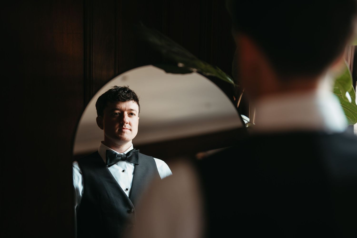 Groom in tuxedo vest and bow tie checking his reflection in a round mirror before wedding ceremony.