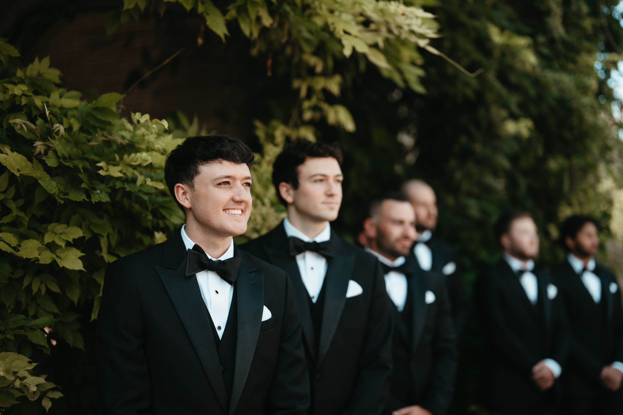 Groomsmen in black tuxedos and bow ties stand outdoors near lush green foliage at a wedding ceremony.