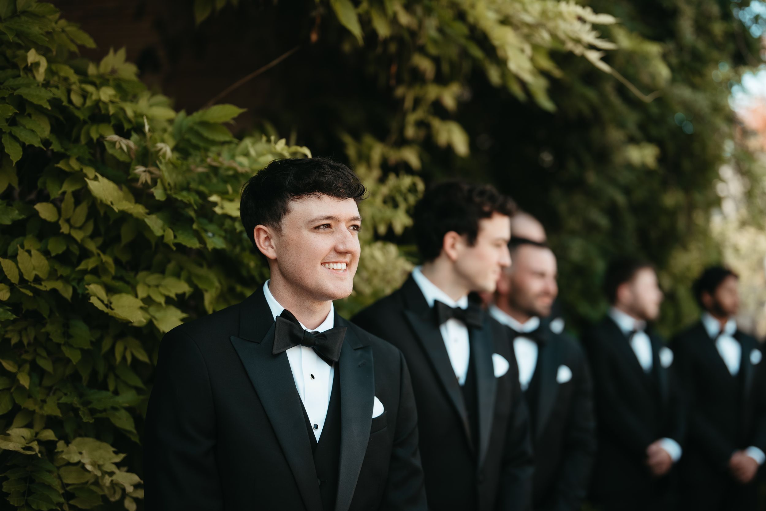 Groom in black tuxedo with bow tie smiling outdoors, groomsmen lined up behind him near lush green foliage.