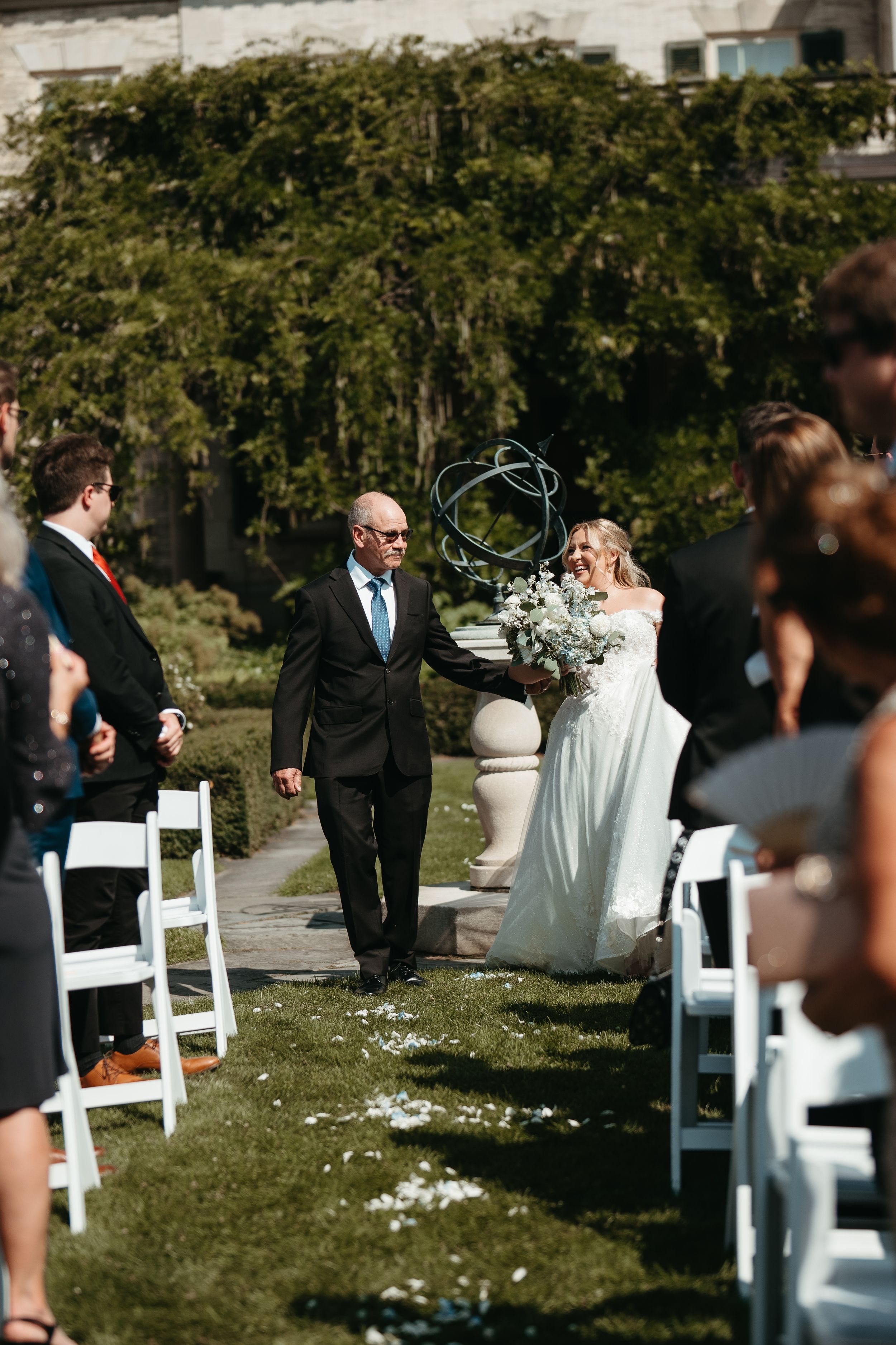 Bride walks down outdoor aisle with escort holding white bouquet at elegant garden wedding ceremony.