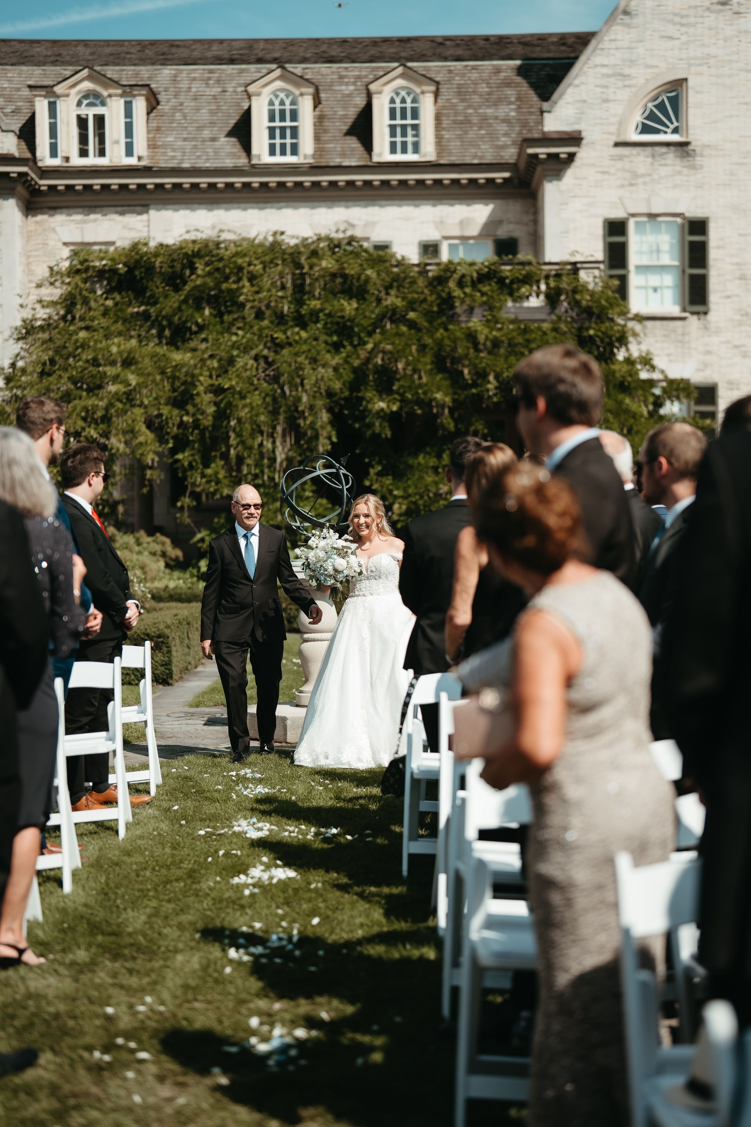Bride and escort walk down aisle at outdoor garden wedding with historic stone mansion backdrop.