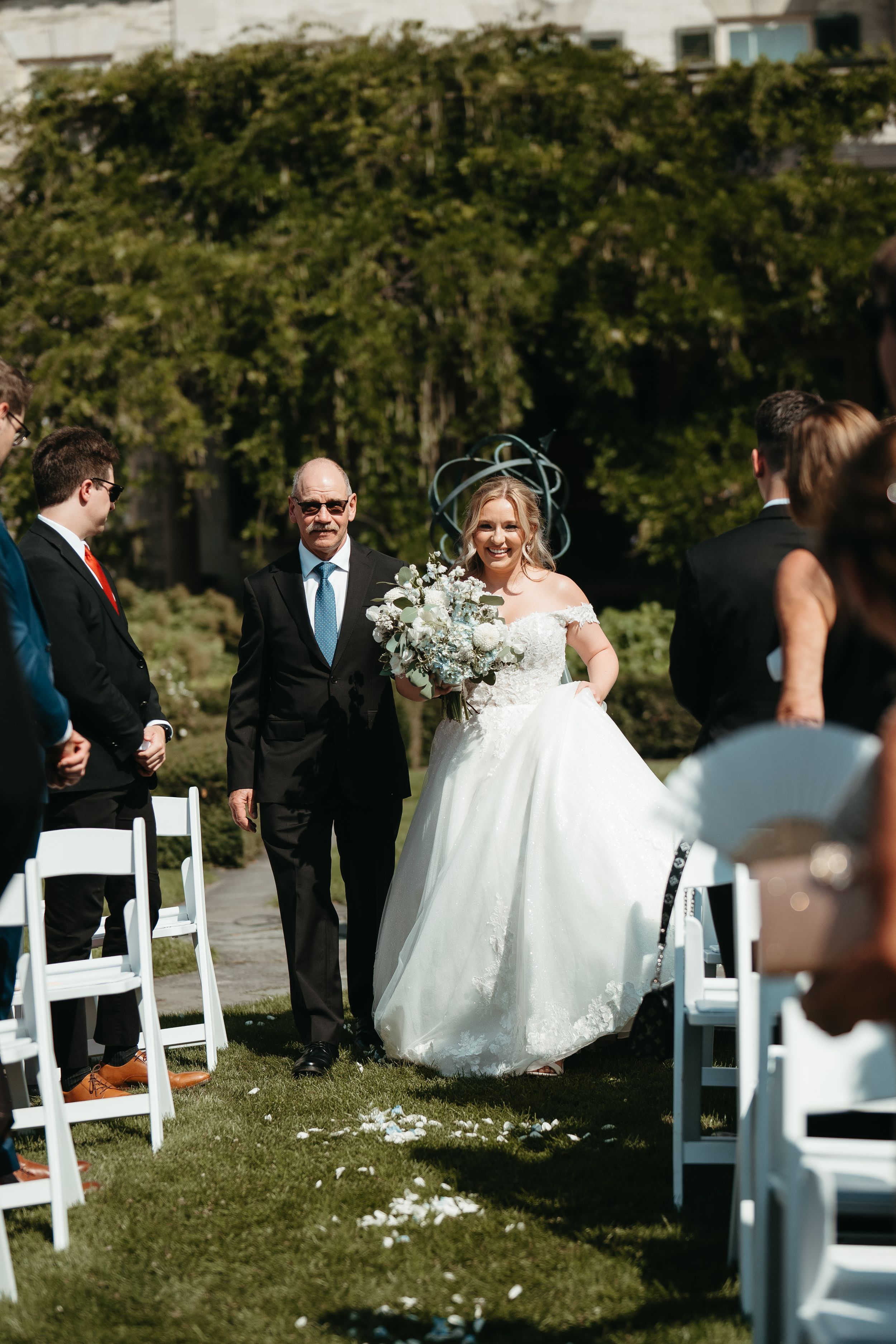Bride in white gown walks down outdoor aisle with escort, holding white bouquet, guests seated in white chairs.