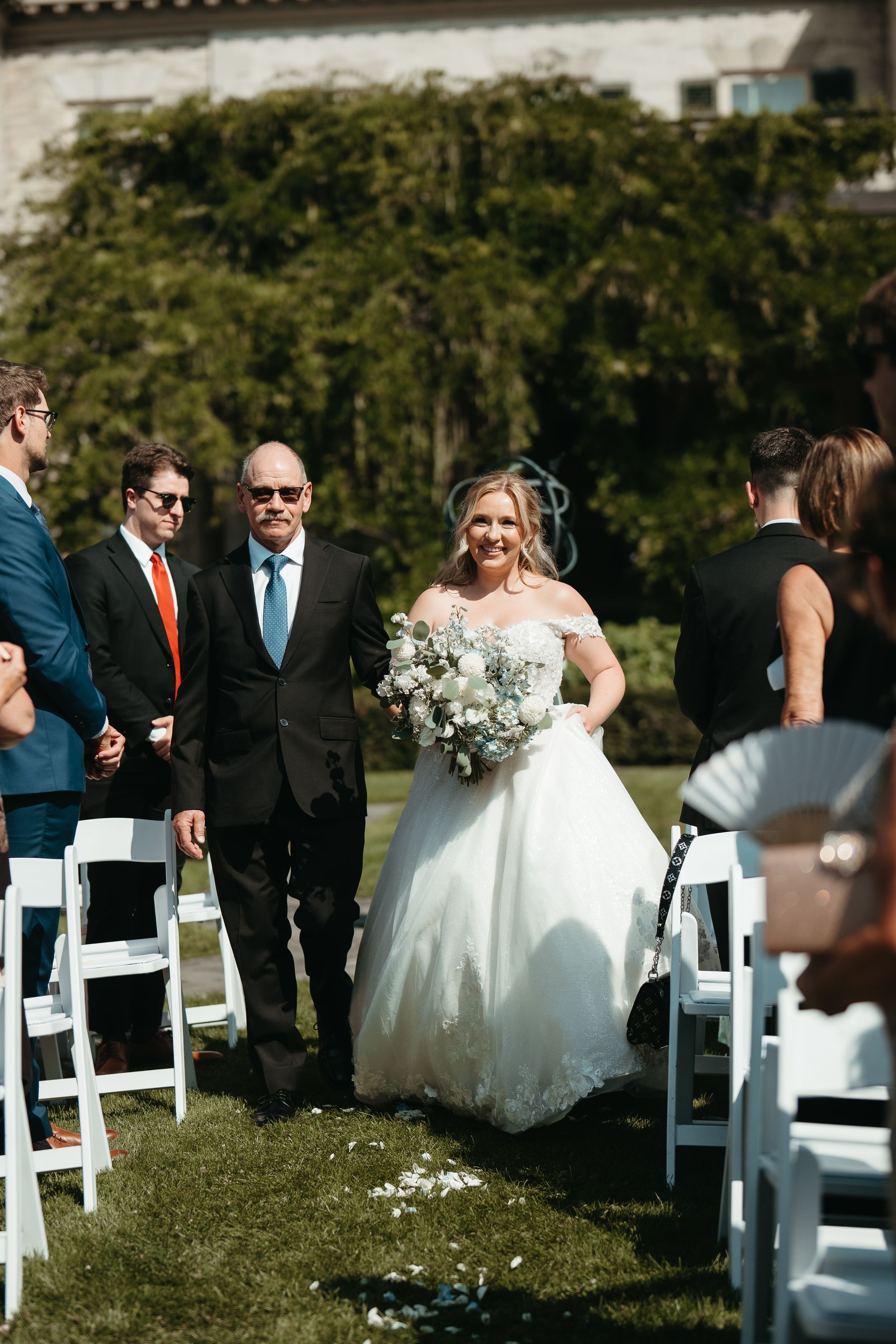 Bride in white off-shoulder gown walks down outdoor aisle with escort, holding white bouquet at sunny wedding ceremony.