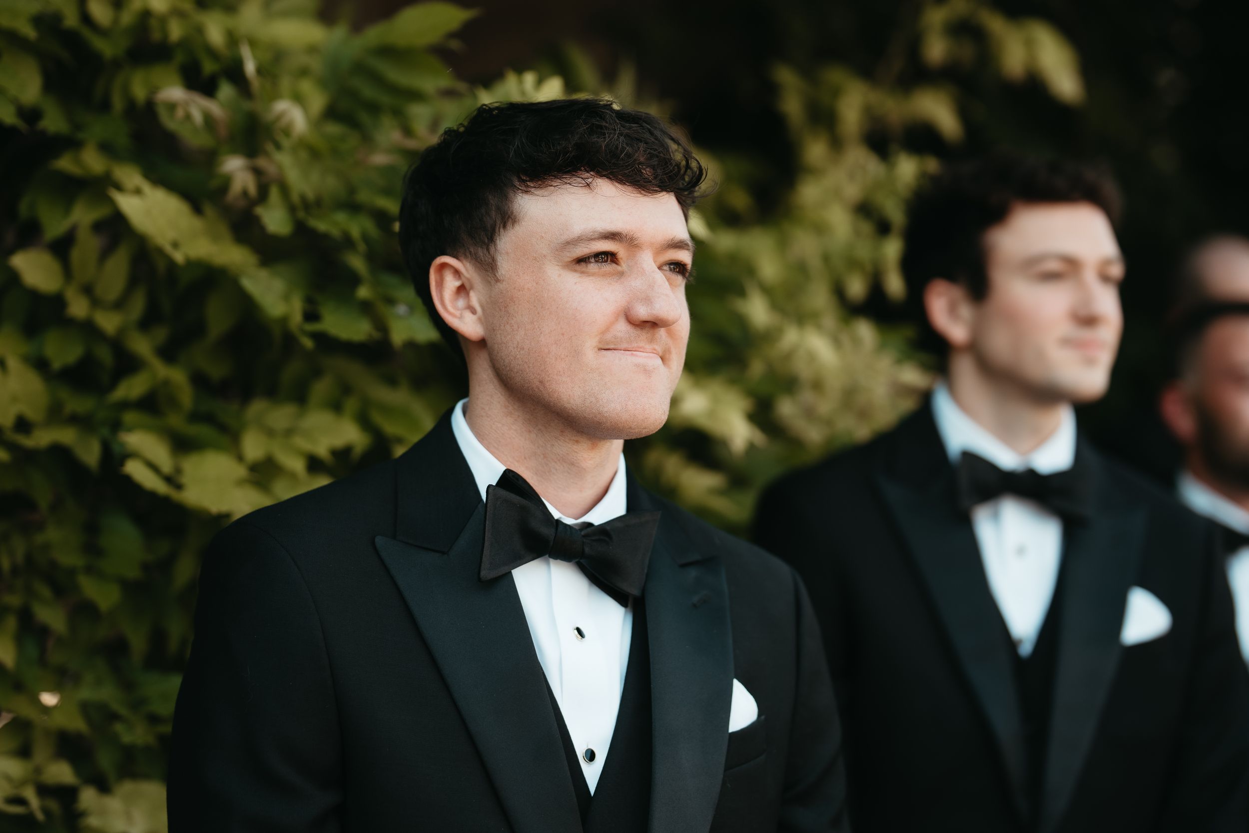 Man in black tuxedo with bow tie smiling outdoors, groomsmen standing in background near green foliage.