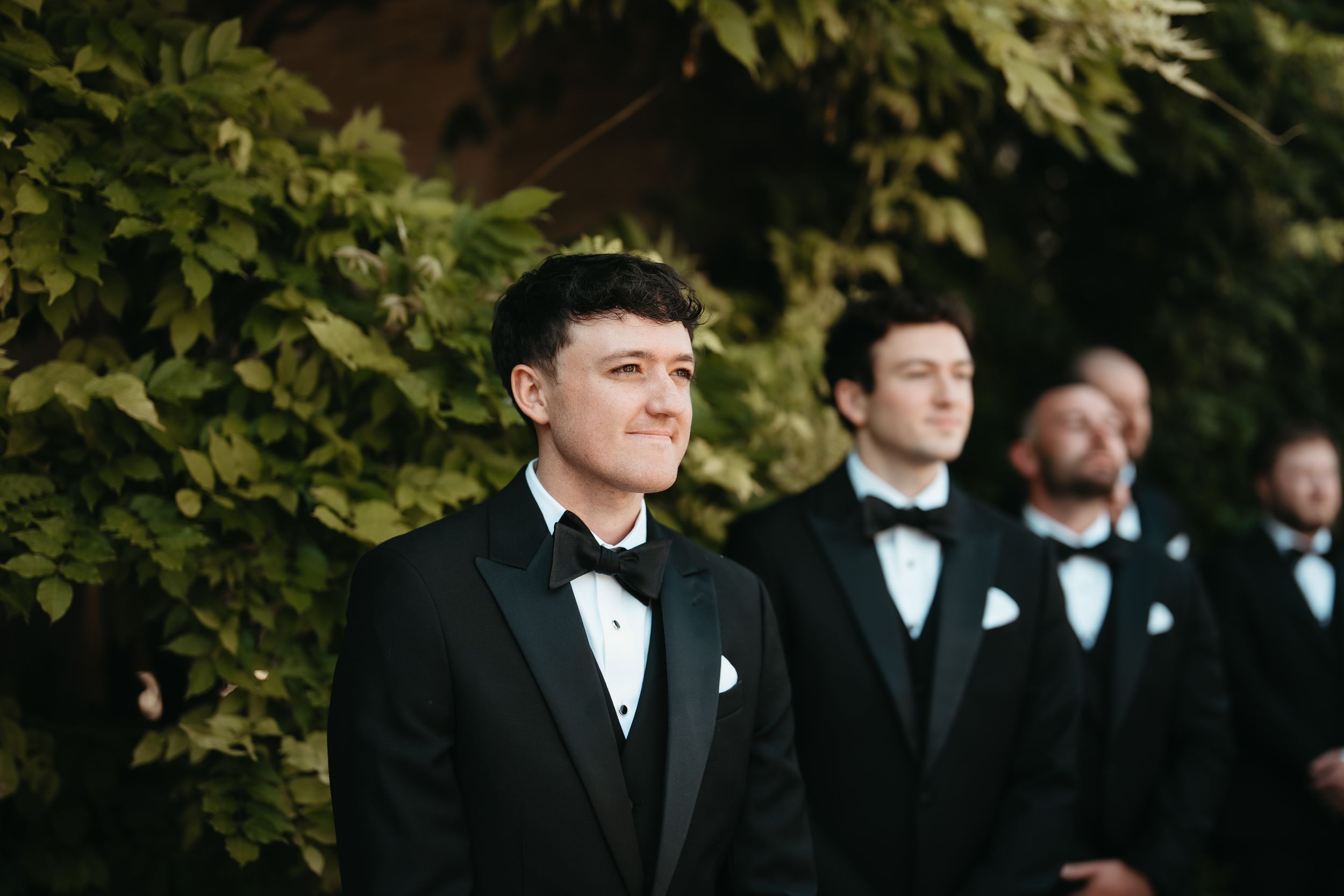 Groom in black tuxedo and bow tie smiling at altar with groomsmen standing behind him at wedding ceremony.