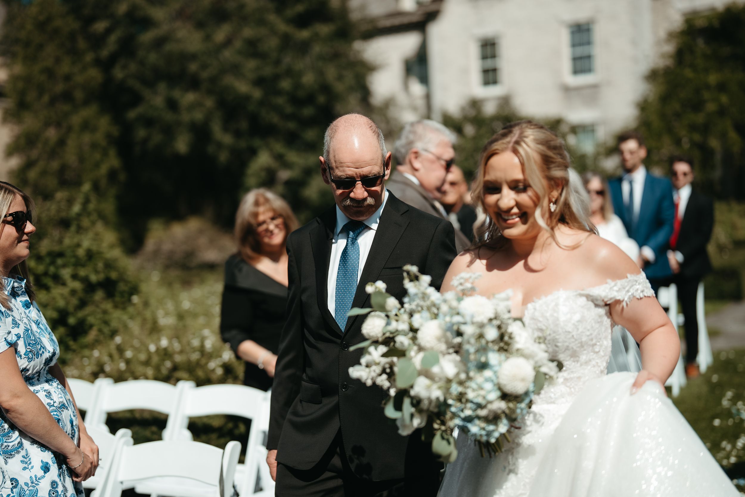 Bride in off-shoulder lace gown holding white bouquet walks down outdoor aisle with escort at sunny wedding ceremony.