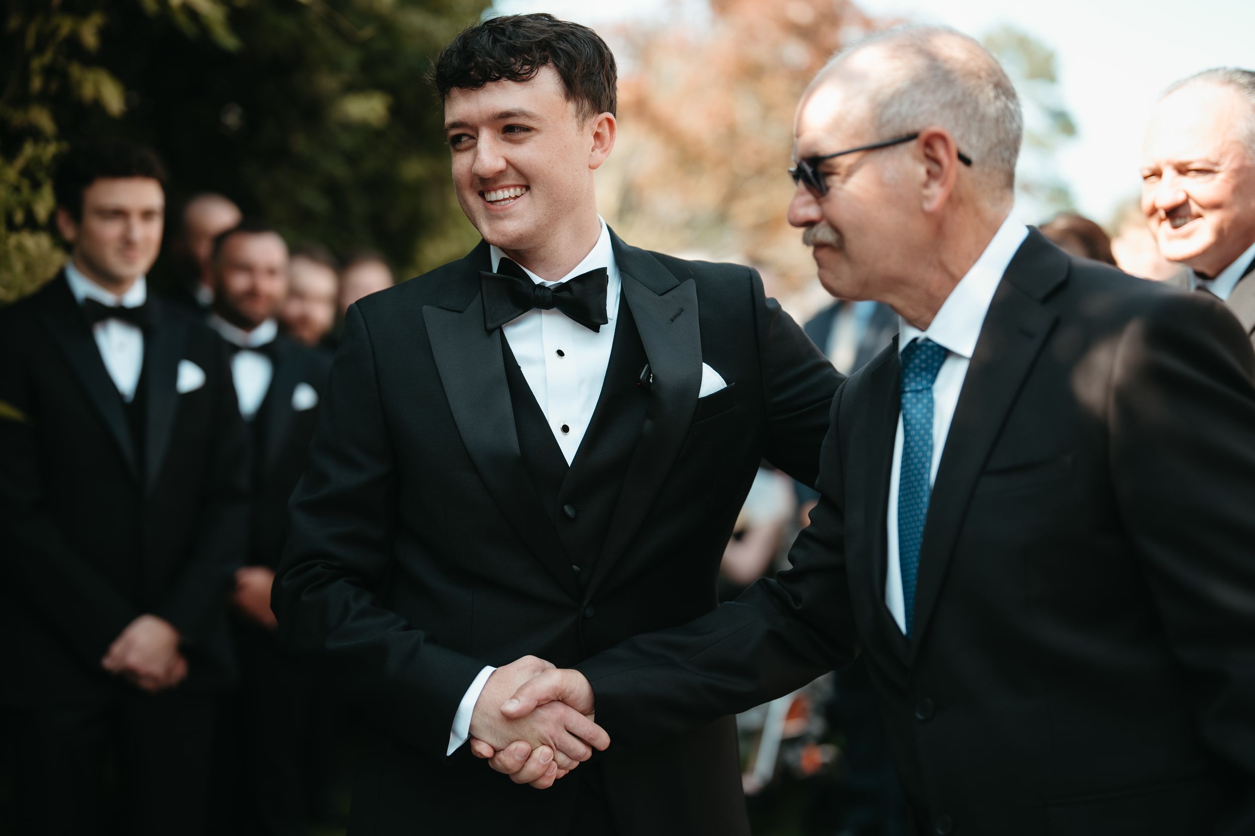 Two men in black tuxedos shaking hands at a formal wedding ceremony outdoors.