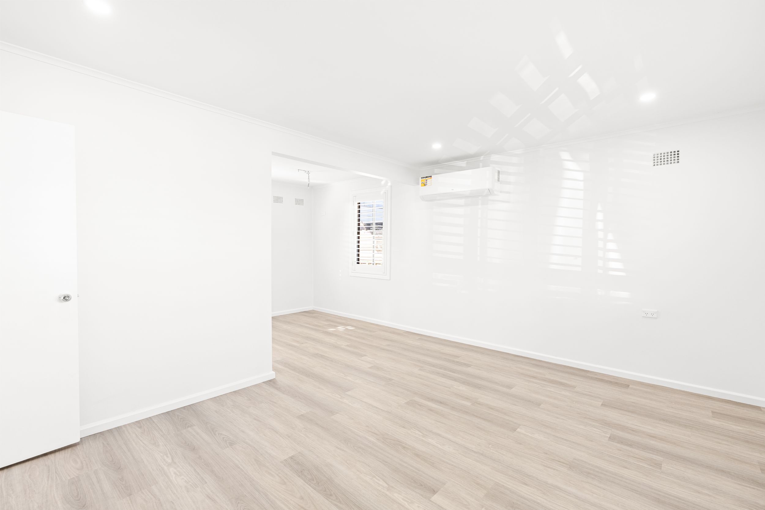Spacious white room with split system air conditioner, plantation shutters, and light timber flooring.