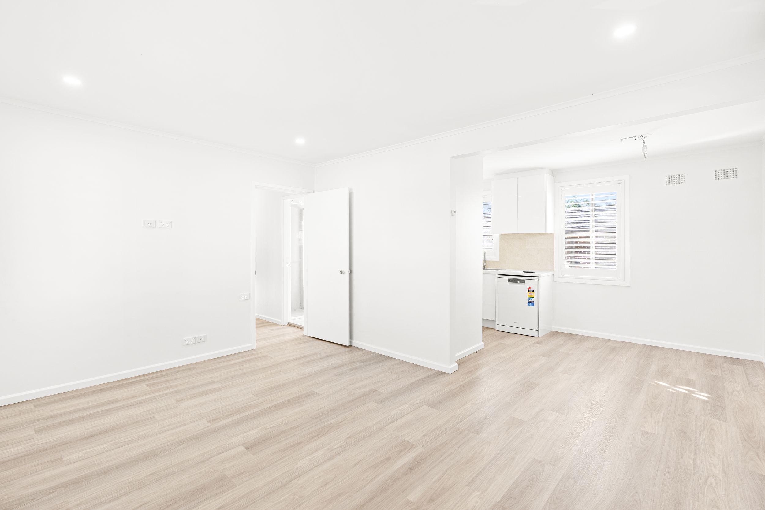 Open plan living area with light timber floors, white walls, and kitchen visible in background.