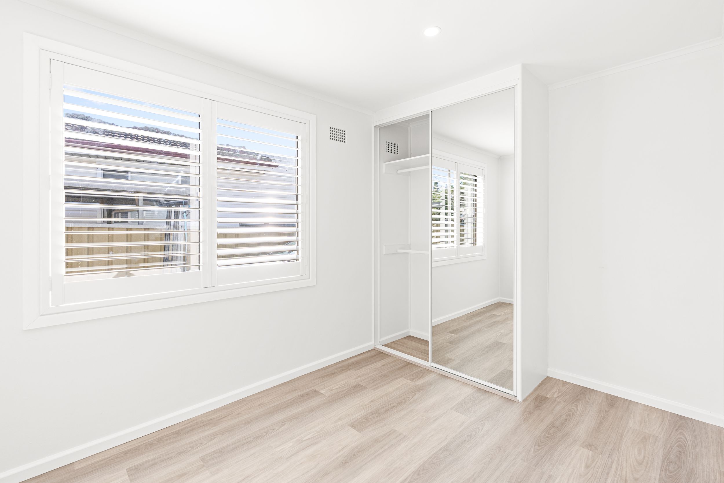 Bright empty bedroom with mirrored built-in wardrobe, plantation shutters, and light timber flooring.