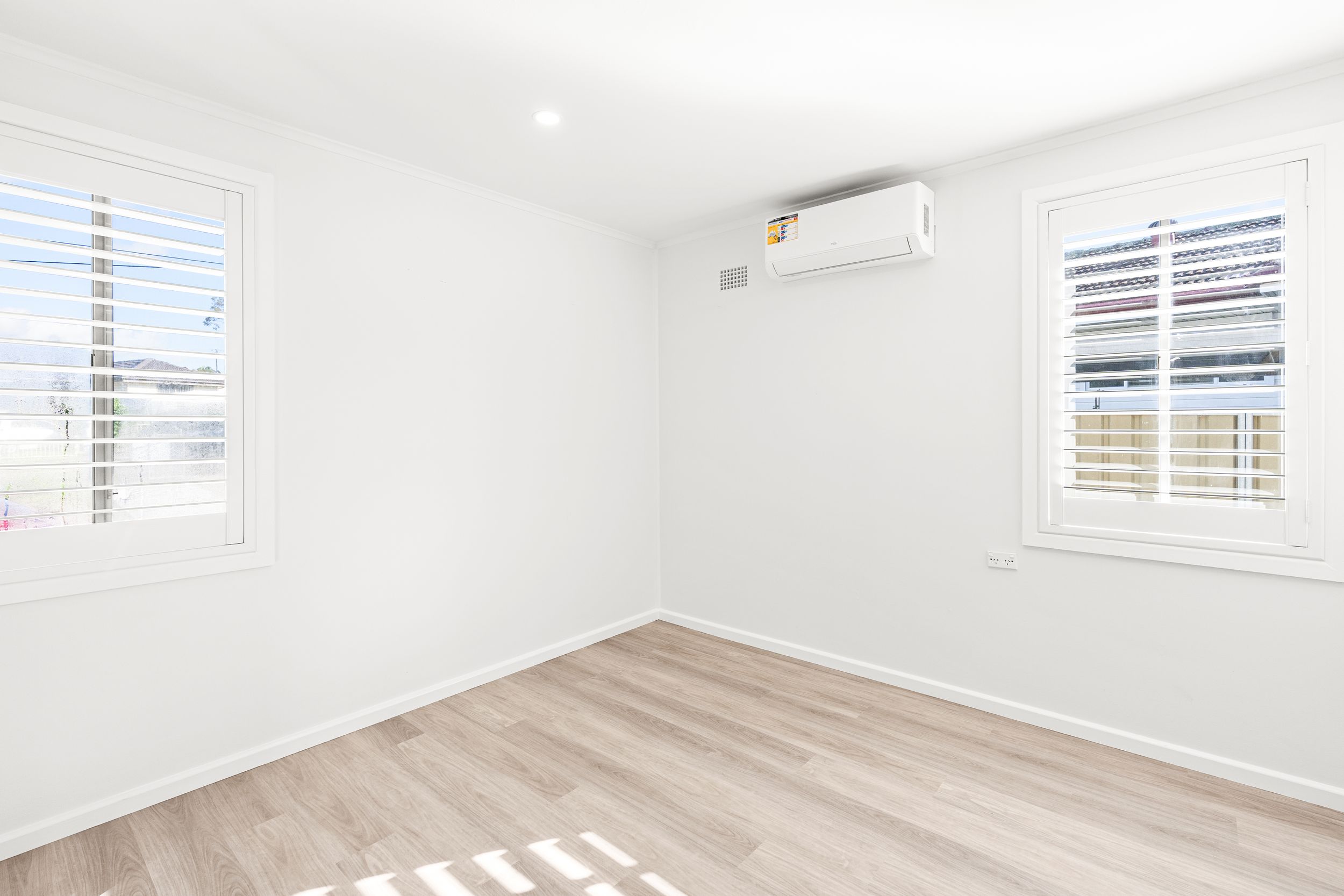 Bright empty room with split system air conditioner, plantation shutters, and light timber flooring.
