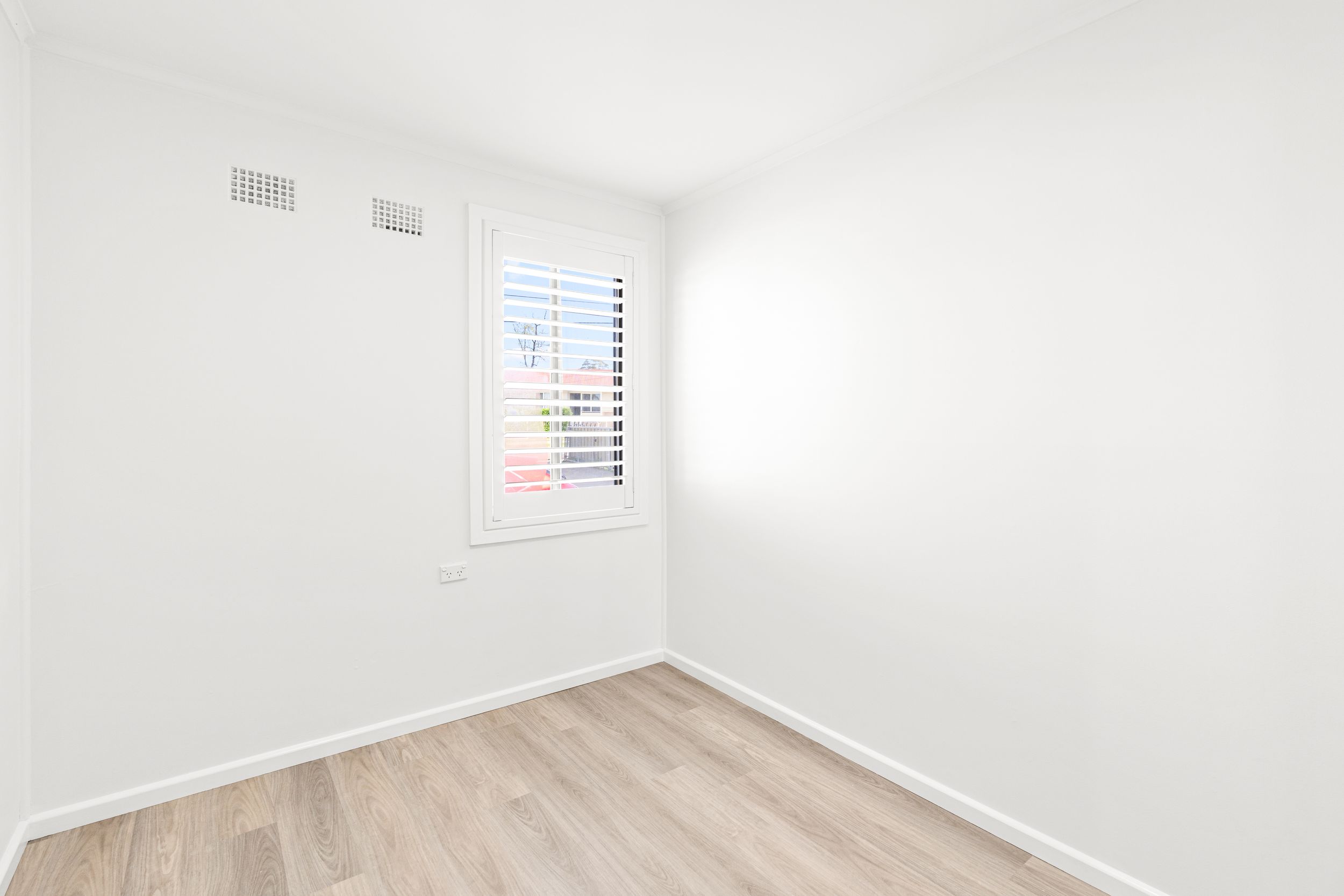 Small bright empty room with plantation shutter window and light timber flooring and white walls.