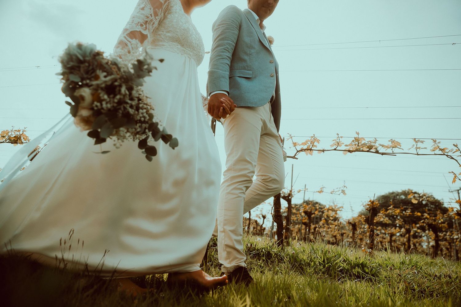 Bride and groom walking hand in hand through vineyard, bride holding bouquet in white gown, groom in light blue suit.
