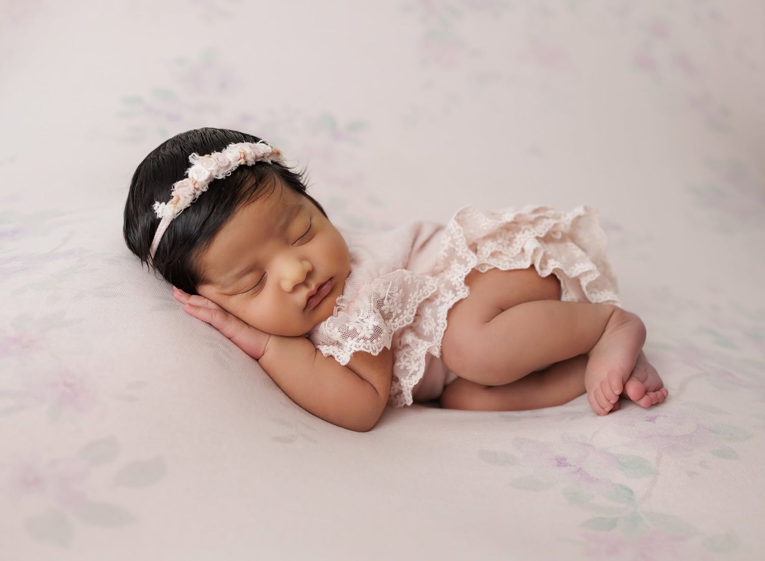 Peaceful newborn baby girl sleeping in pink lace romper and floral headband on white fluffy blanket.