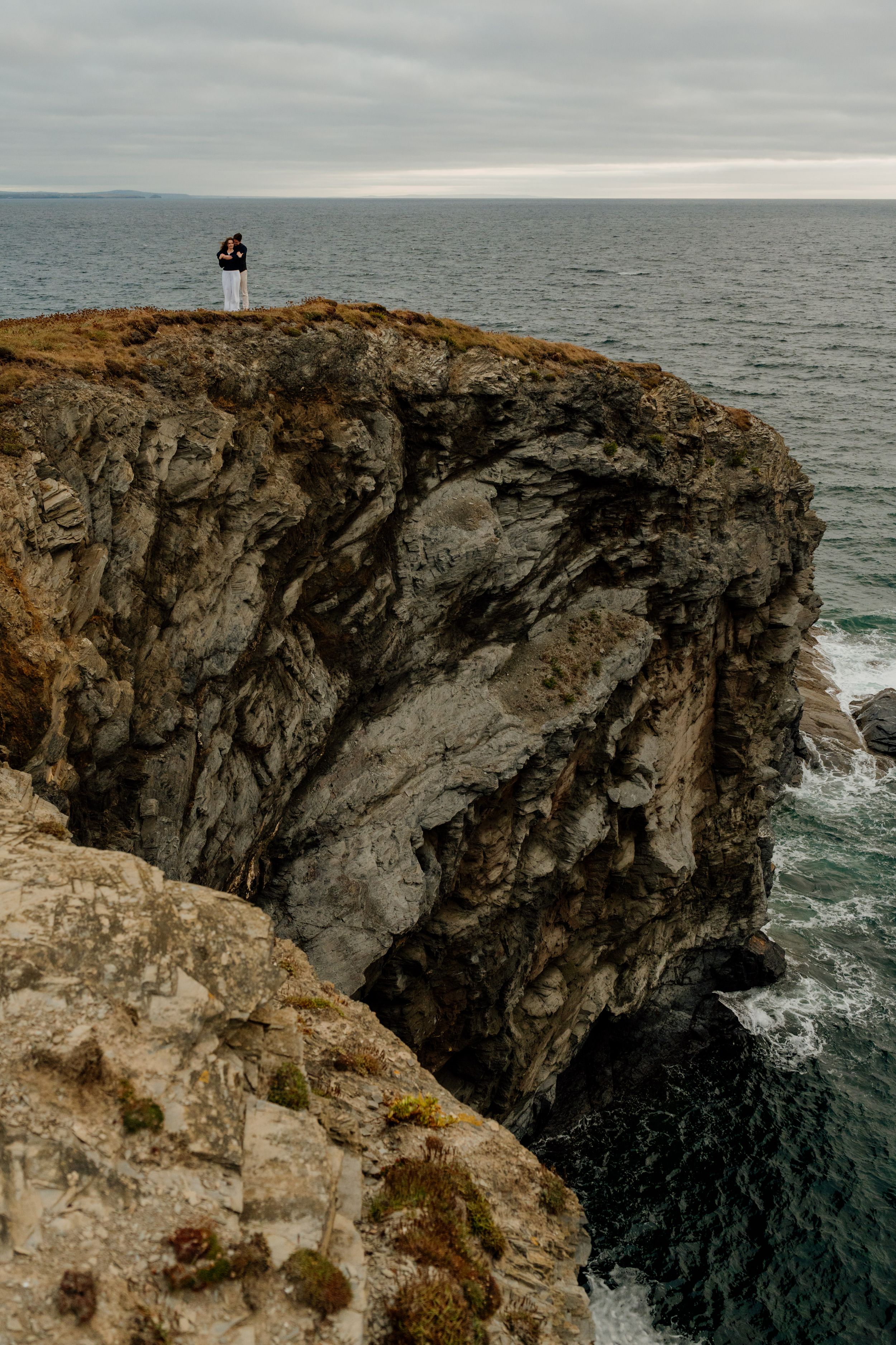 Couple standing on dramatic rocky coastal cliffs overlooking the vast ocean under overcast skies.