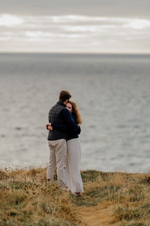 Couple embracing on coastal cliff overlooking calm ocean waters on an overcast day, wearing casual autumn attire.