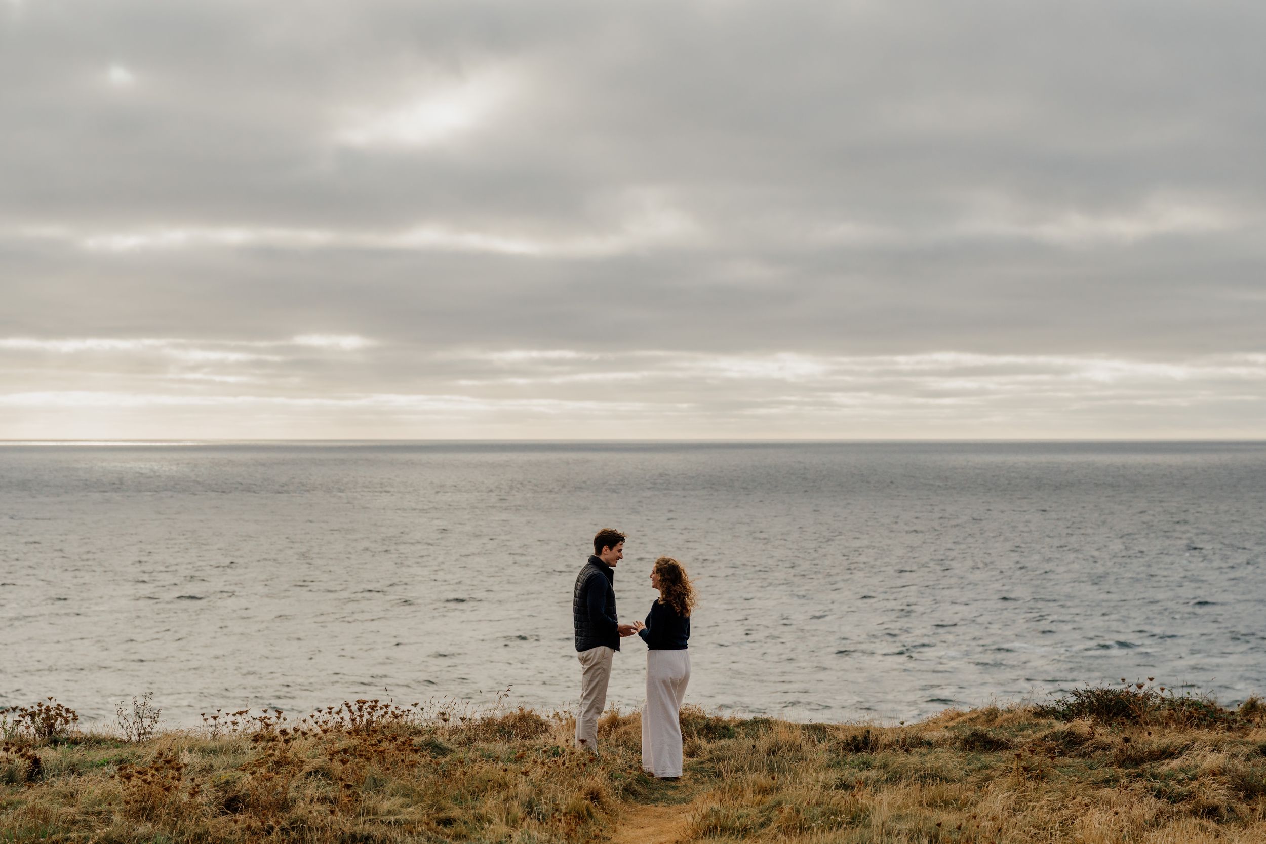 Couple holding hands on coastal cliffside with overcast sky and ocean backdrop during romantic engagement photo session.