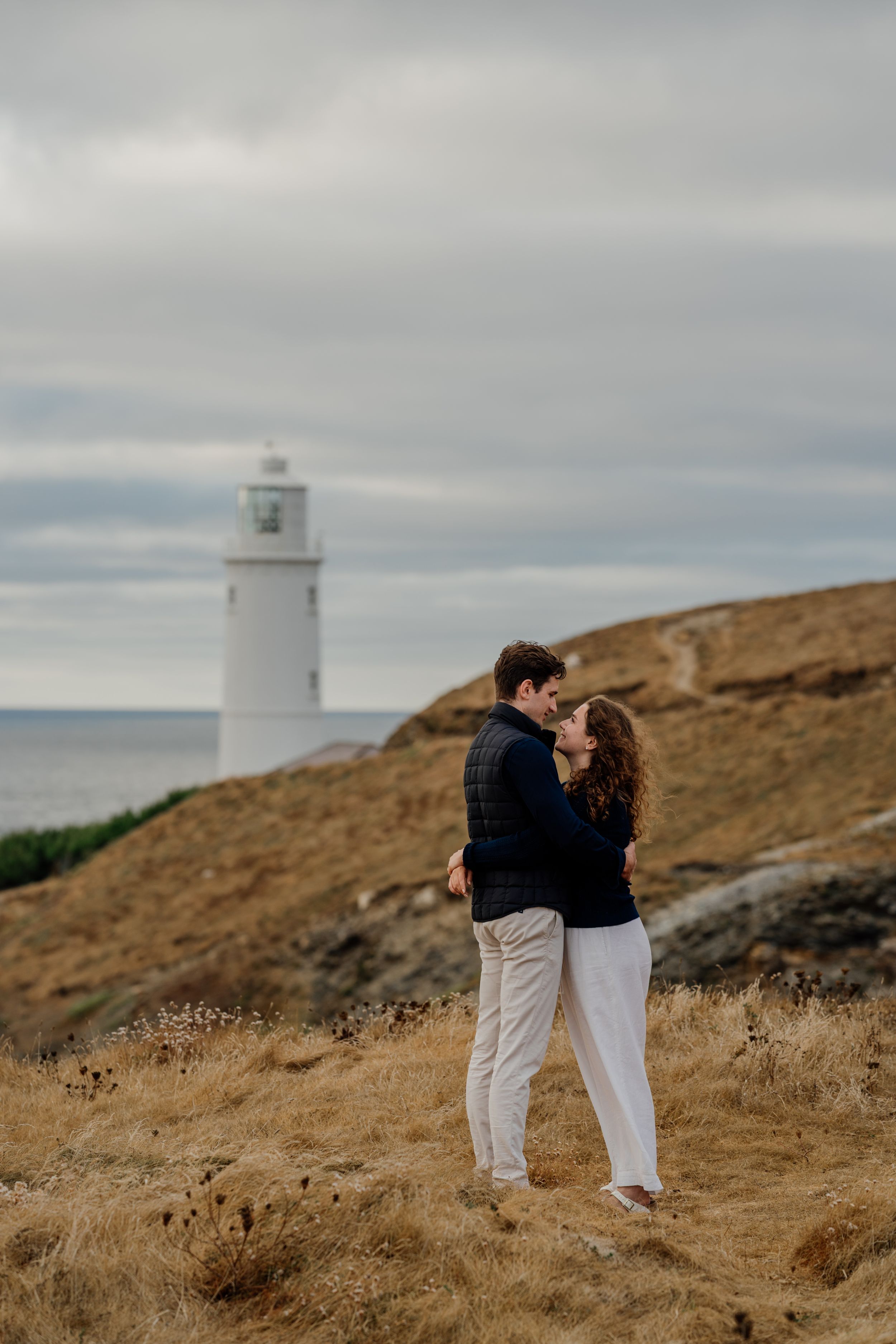 Couple embracing on a grassy coastal hillside with a white lighthouse and ocean in the background.
