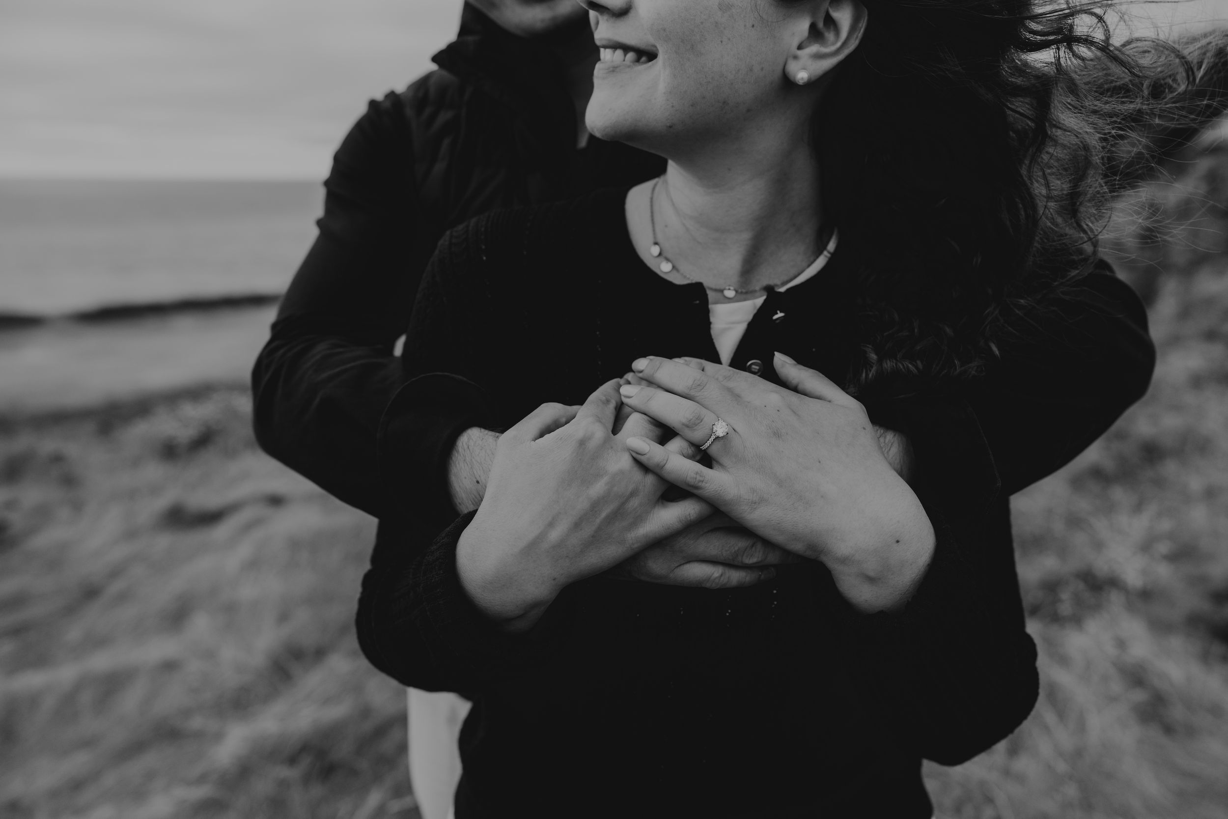 Black and white photo of couple embracing on beach, woman smiling, showing engagement ring, ocean in background.