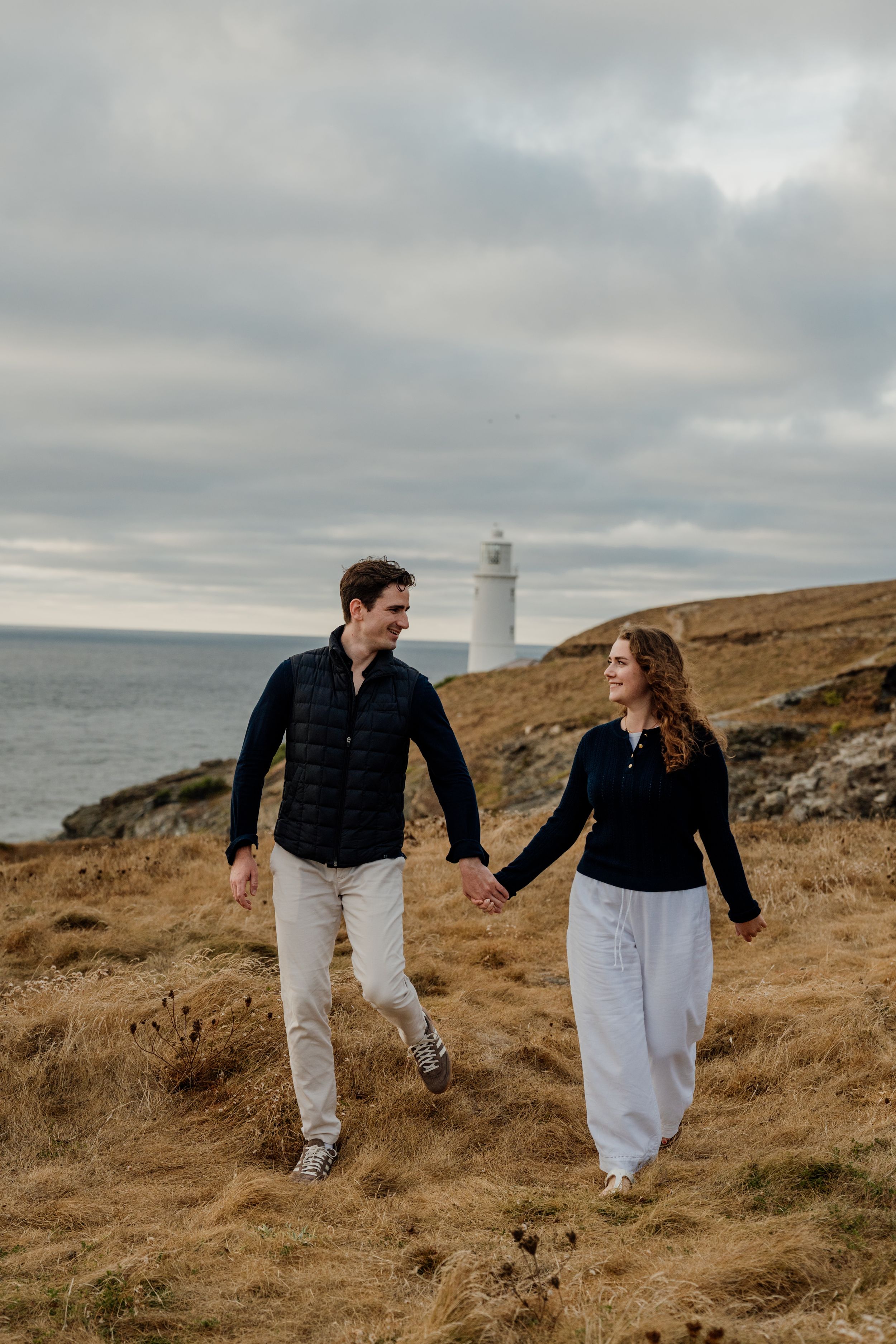 Couple holding hands walking through coastal grassland with white lighthouse and cloudy sky in background.
