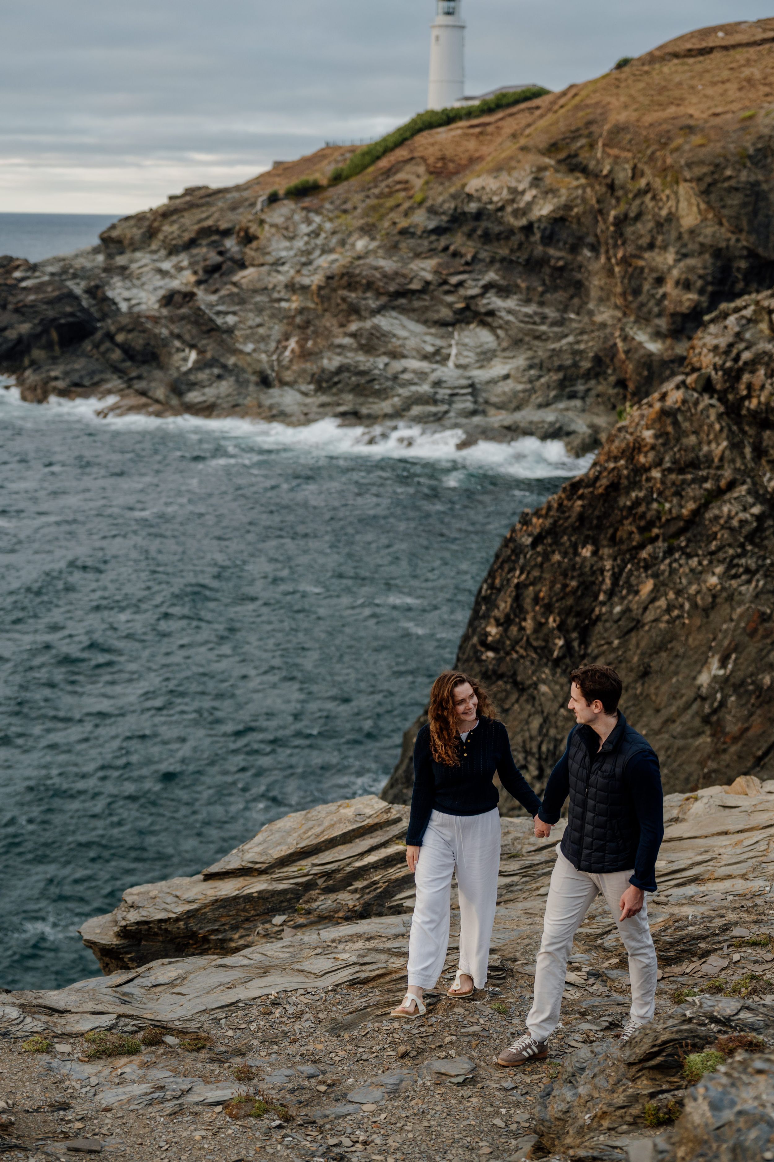 Couple holding hands on rocky coastal cliffs with crashing waves and lighthouse perched above them.