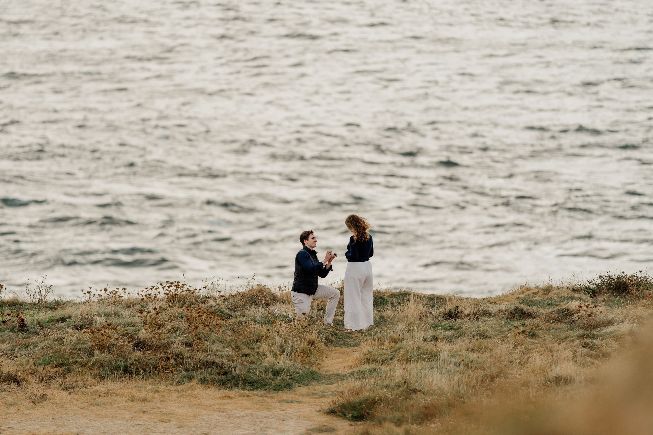 Couple sharing a romantic proposal moment on a coastal cliffside overlooking the ocean.
