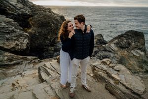 Couple embracing on rocky coastal cliffs, wearing navy sweaters and white pants, smiling with ocean waves in background.
