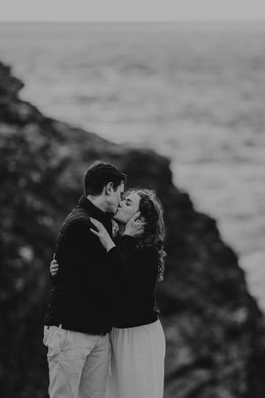Black and white photo of a couple sharing a romantic kiss on rocky coastal cliffs with ocean waves in the background.