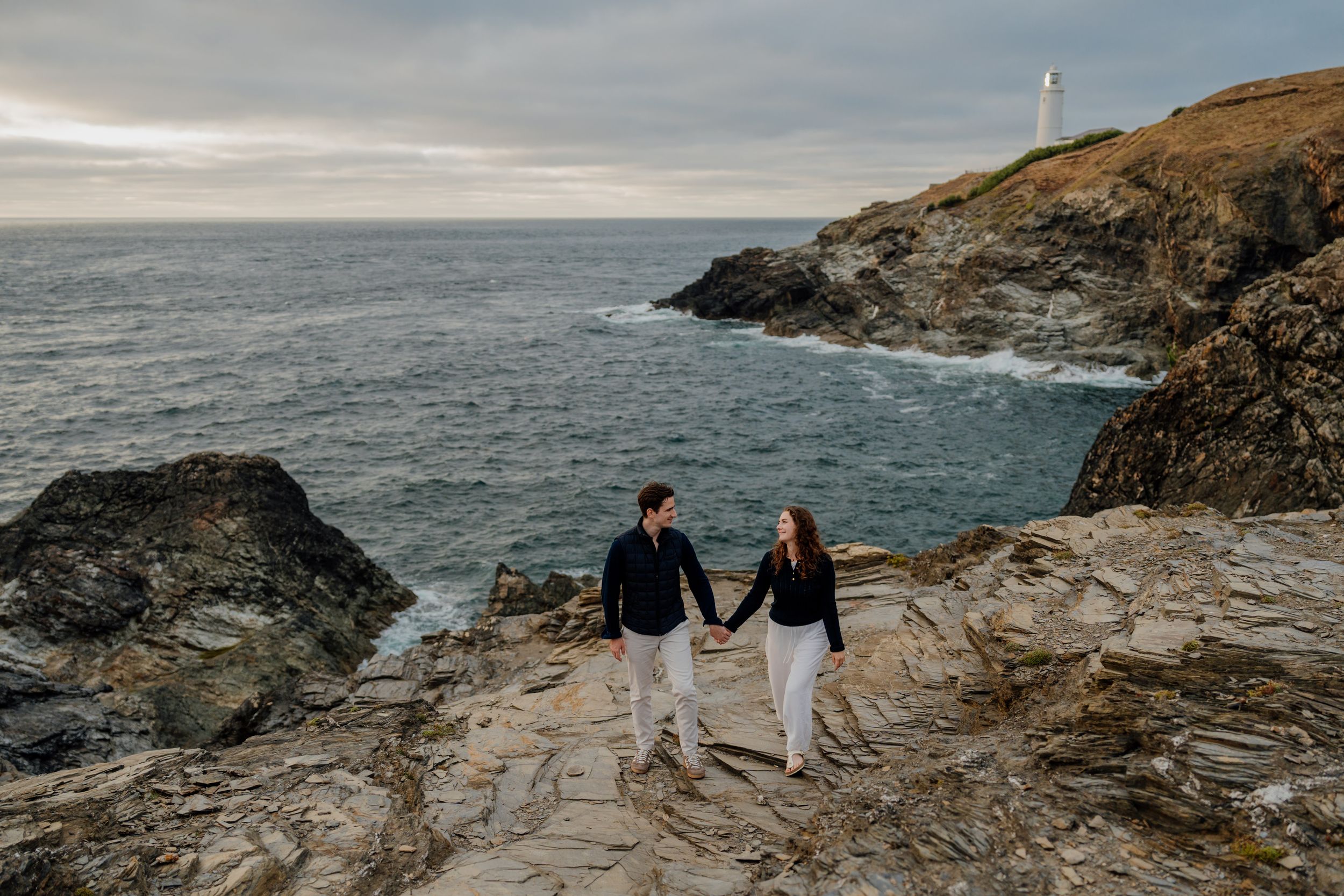 Couple holding hands on rocky coastal cliffs with a lighthouse and ocean waves in the background.