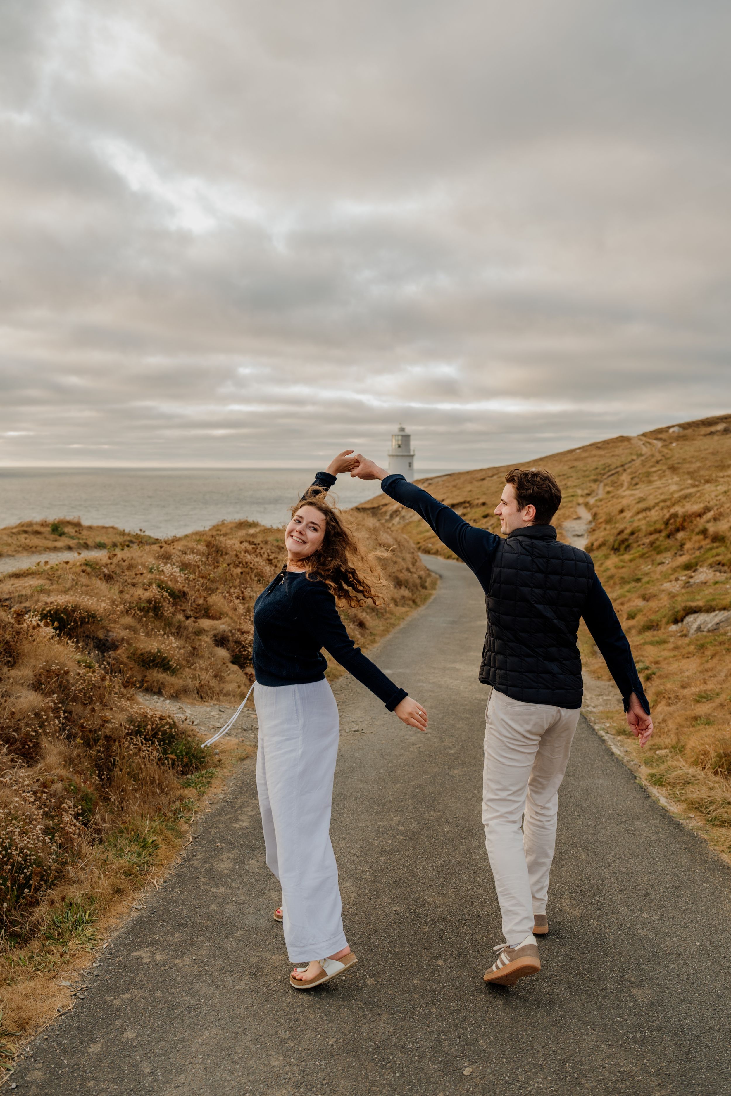 Couple dancing on coastal road near lighthouse, woman in white pants spinning, dramatic cloudy sky and ocean backdrop.