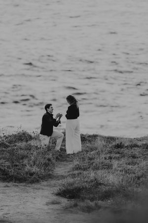 Black and white photo of a man on one knee proposing to a woman on a coastal cliffside path.