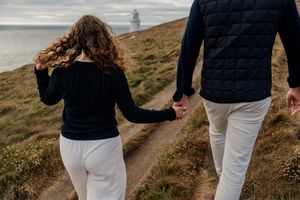 Couple holding hands walking on a coastal path toward a lighthouse on a cloudy day.