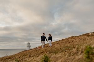 Couple walking hand in hand on a grassy hillside with a lighthouse and cloudy sky behind them.