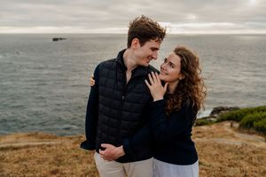 Couple embracing and smiling at each other on a coastal cliff with ocean views on a cloudy day.