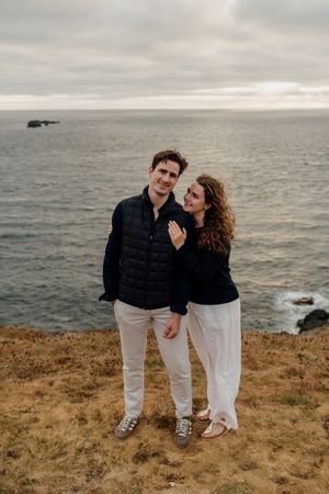 Couple posing on coastal cliffside with ocean backdrop, wearing matching dark tops and white pants.