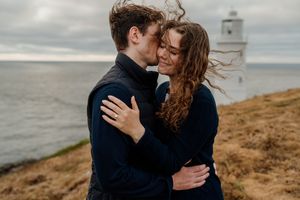 Couple embracing near a lighthouse on a coastal cliff, sharing a romantic moment with wind-swept hair.