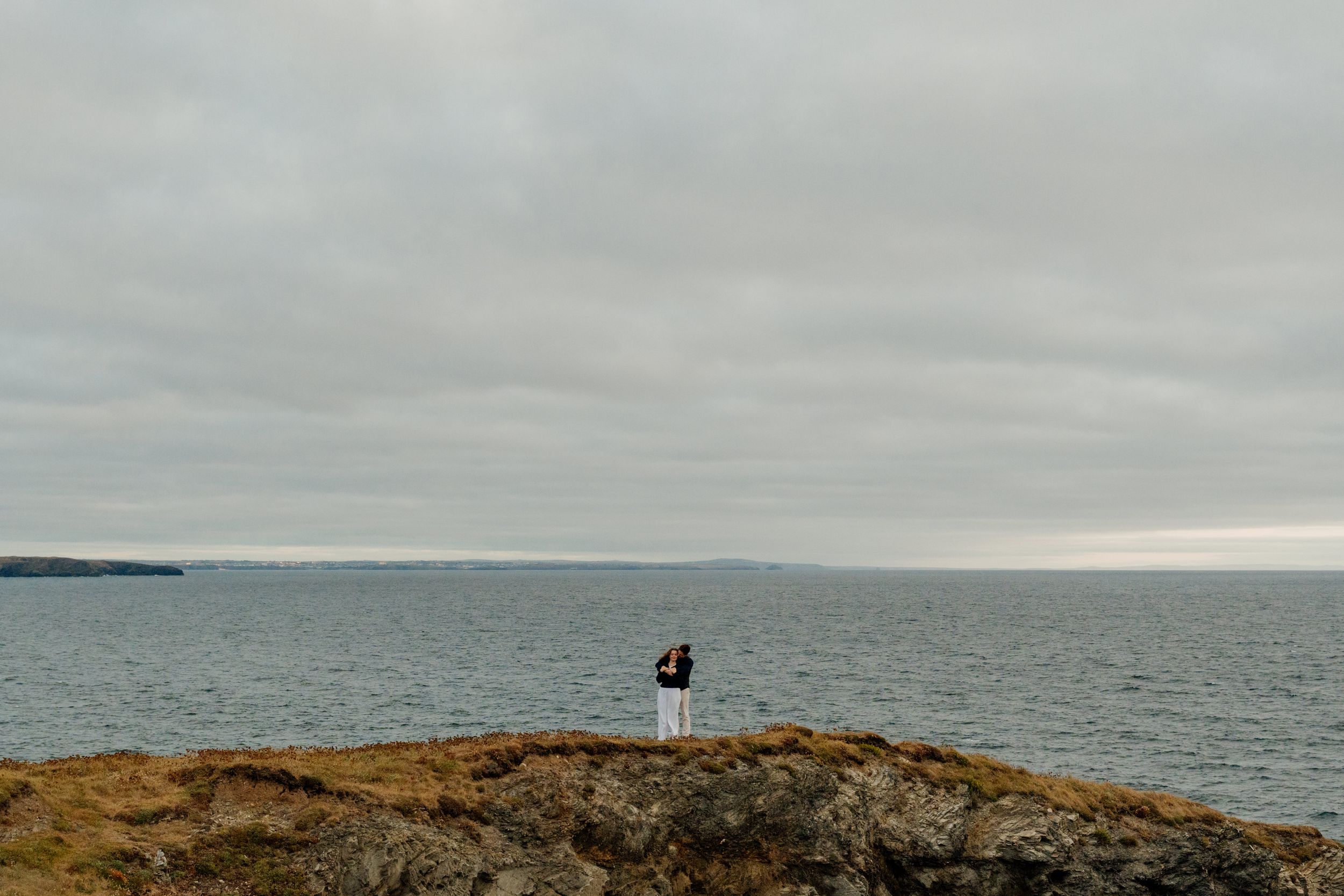 A lone figure stands on rocky coastal cliffs overlooking a vast grey ocean under an overcast sky.