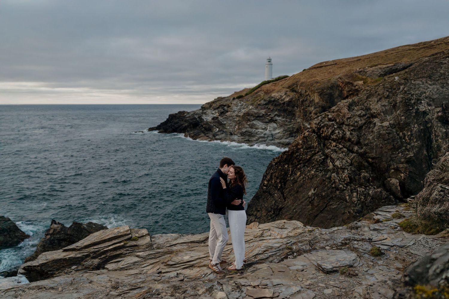 Couple embracing on rocky coastal cliffs with ocean waves and trevose head lighthouse in the background under overcast skies.