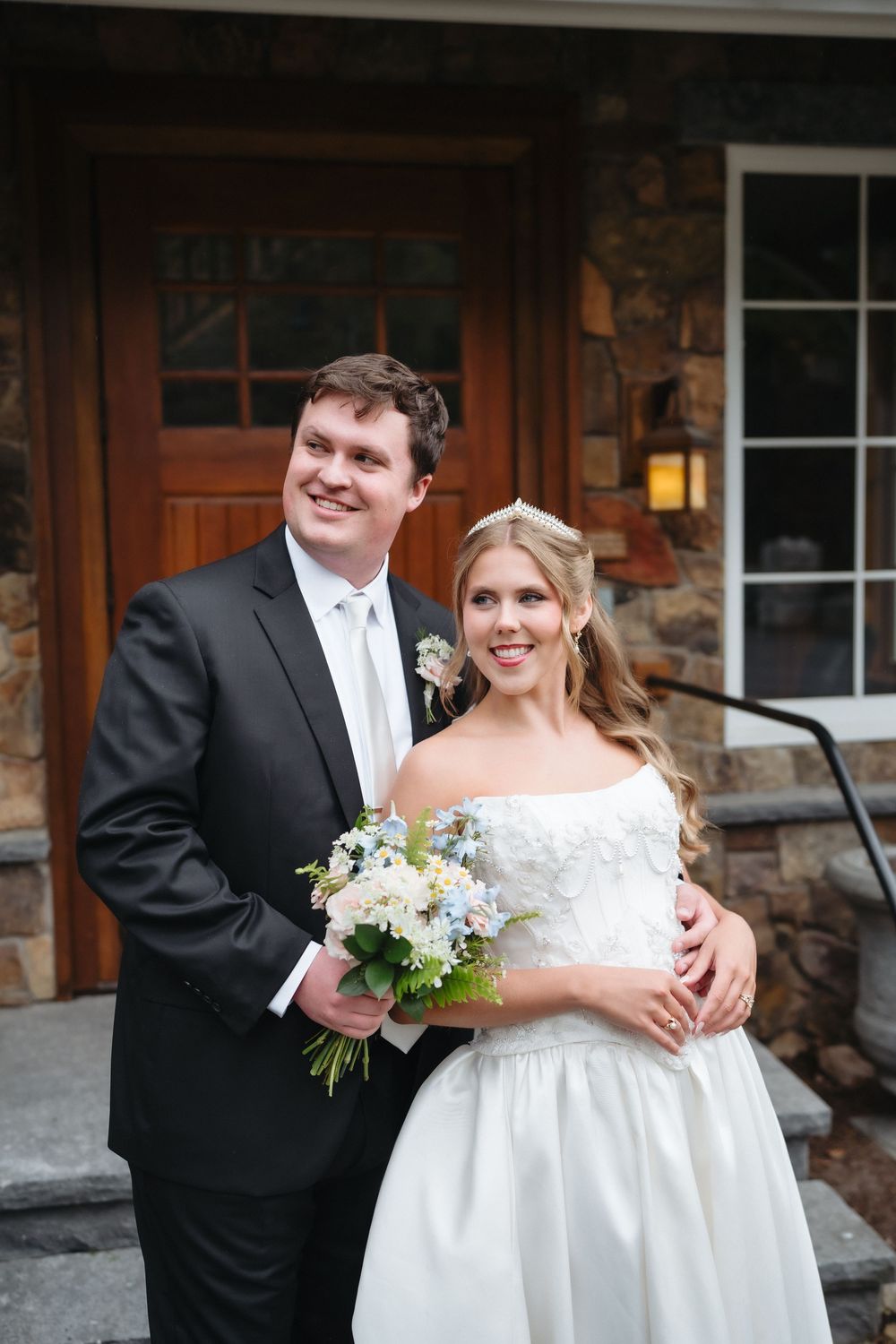 Smiling bride and groom pose together outside stone building, bride holding blue and white bouquet in white gown.