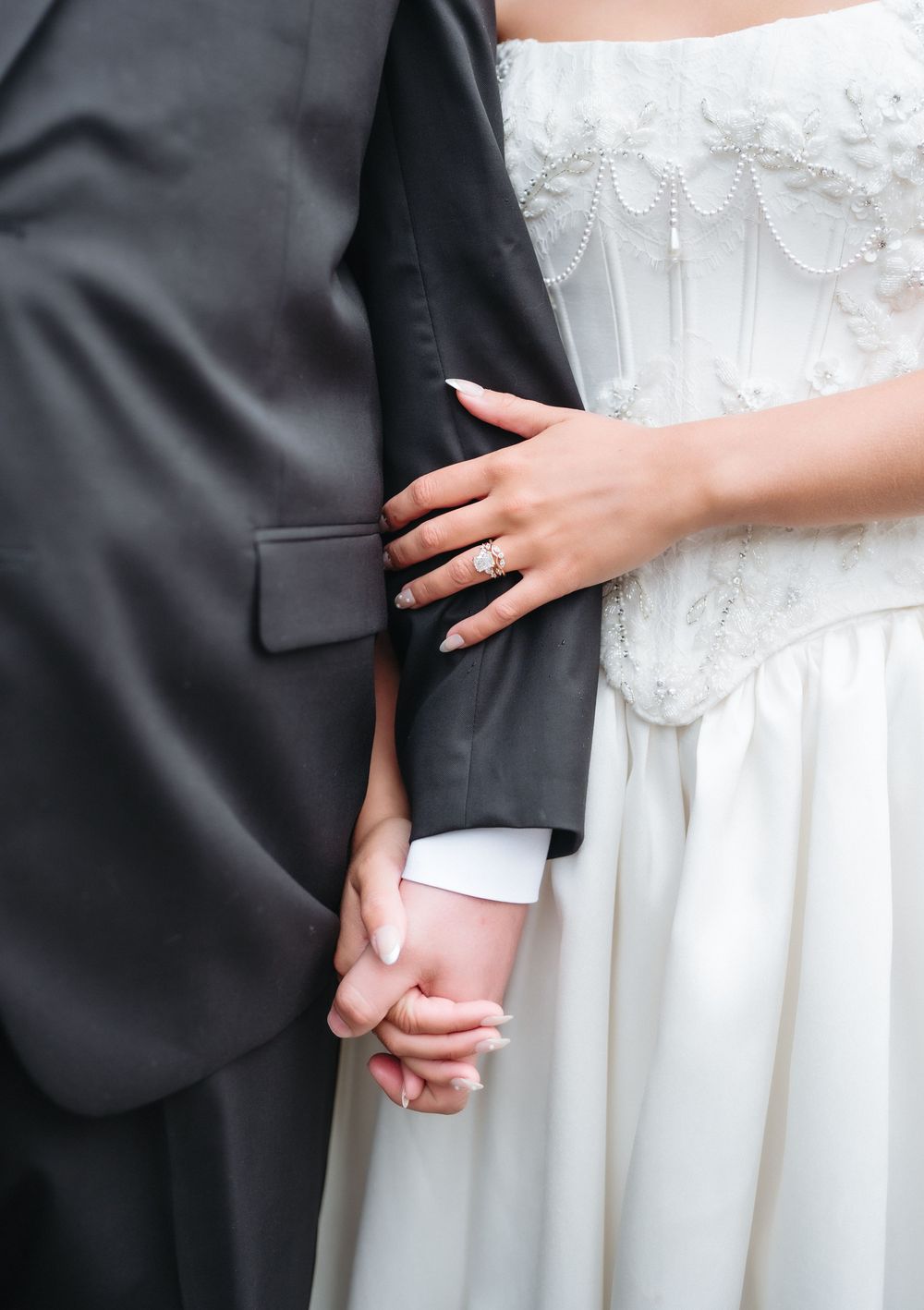 Bride in white lace gown holds groom's arm in black suit, showing diamond engagement ring and manicured nails.