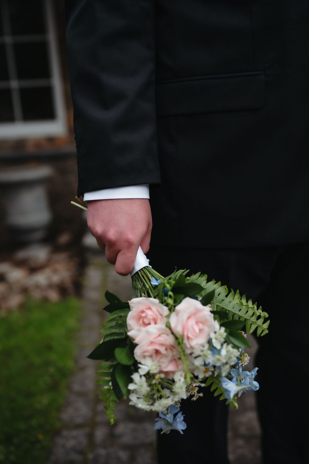 Groom in black suit holding a bridal bouquet of pink roses, white flowers, and greenery at a wedding.